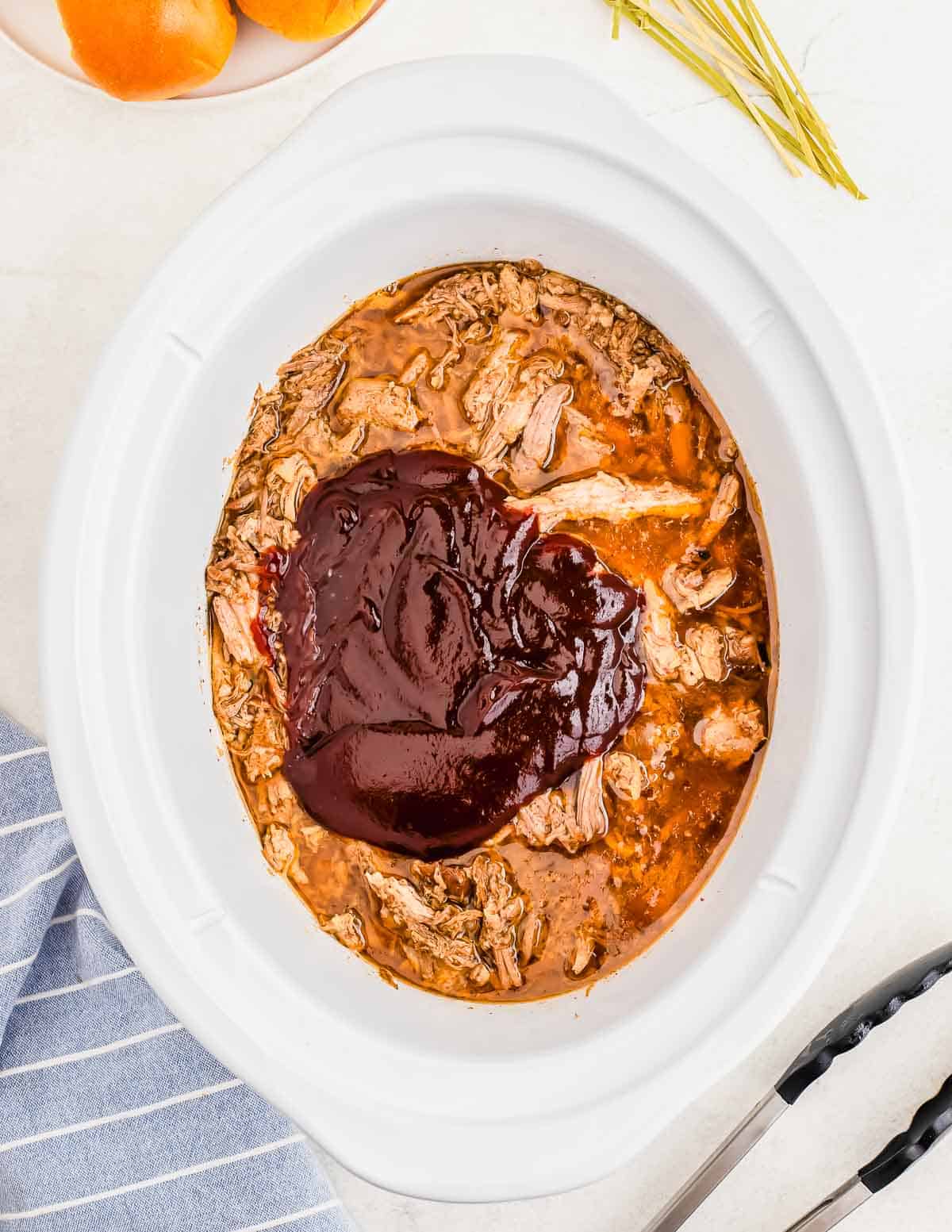 A white slow cooker filled with shredded pork topped with a generous amount of barbecue sauce. Nearby are a kitchen towel, tongs, and a plate with sliced bread.