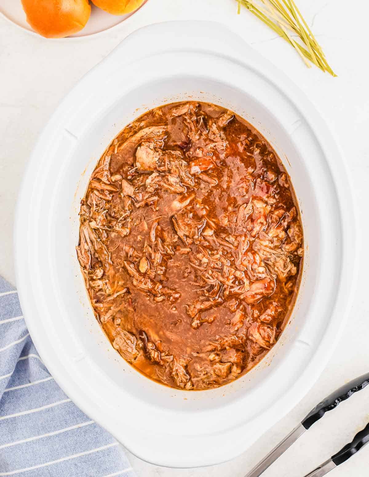 A slow cooker filled with pulled pork in a BBQ sauce. White surface with a striped cloth, tongs, and a bowl of oranges in the background.