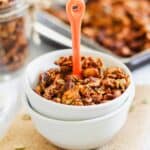 Bowl of homemade granola with nuts and pumpkin seeds, served with an orange spoon, placed on a burlap mat. A jar of granola and a tray in the background.