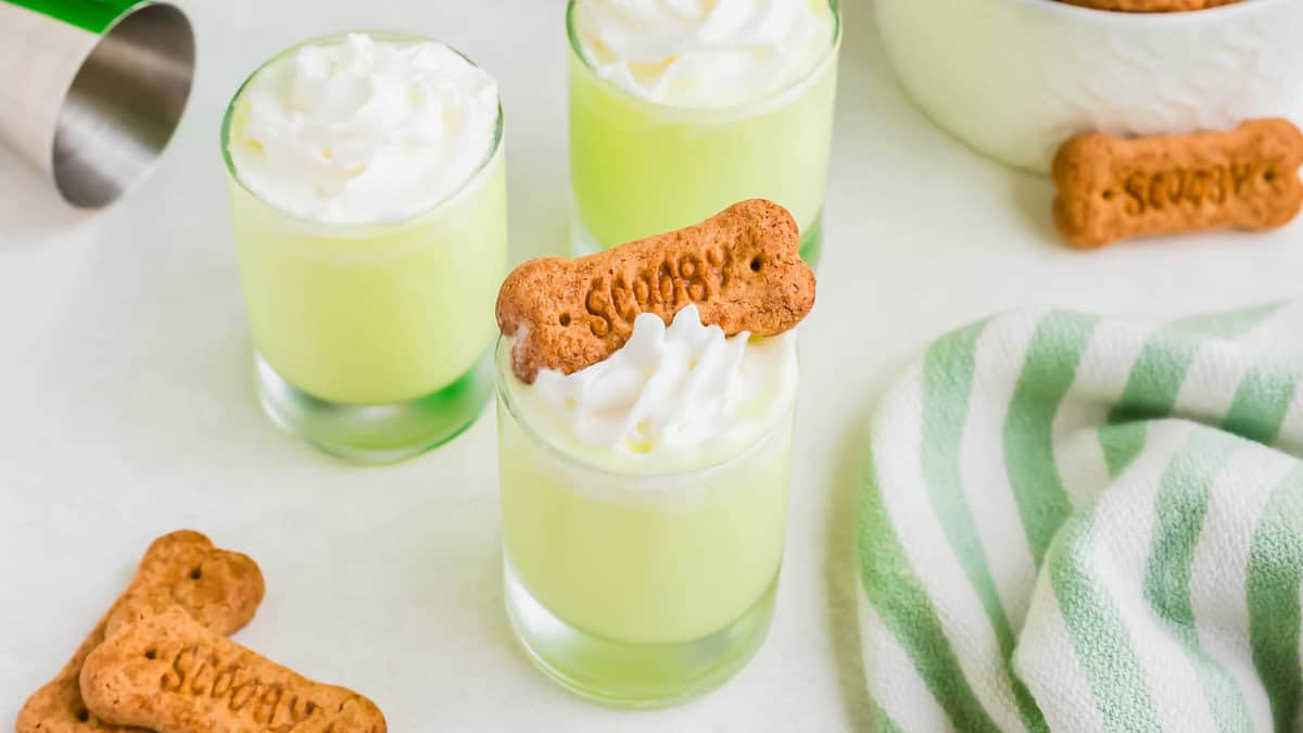 Three glasses of a light green Scooby Snack shot, topped with whipped cream and a cookie.