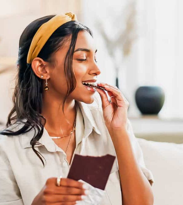 A woman with a yellow headband sits on a couch and eats a piece of chocolate. She is holding another piece of chocolate in her left hand.