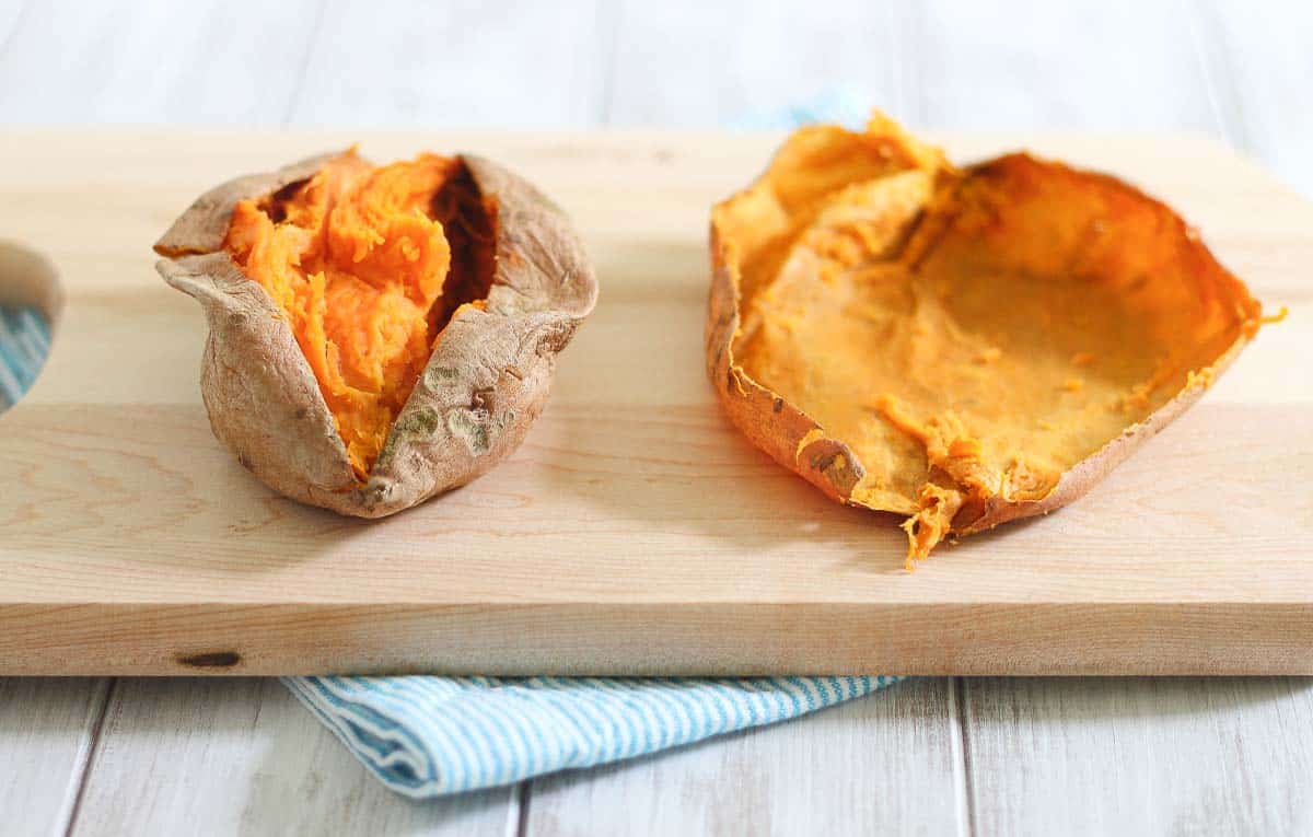 A split baked sweet potato, with the flesh scooped out, lies on a wooden cutting board with a blue-striped cloth underneath.