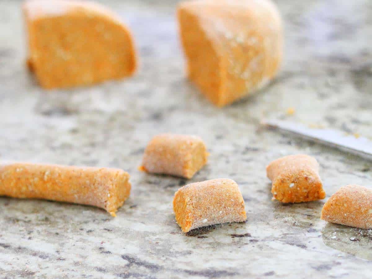 Close-up of raw gnocchi dough pieces being cut on a marble countertop, with some rolled dough and a knife visible in the background.