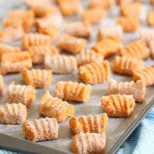 A baking tray filled with uncooked homemade sweet potato gnocchi, lightly dusted with flour. The gnocchi pieces are evenly spaced and have a ridged texture. The tray is on a striped blue cloth.