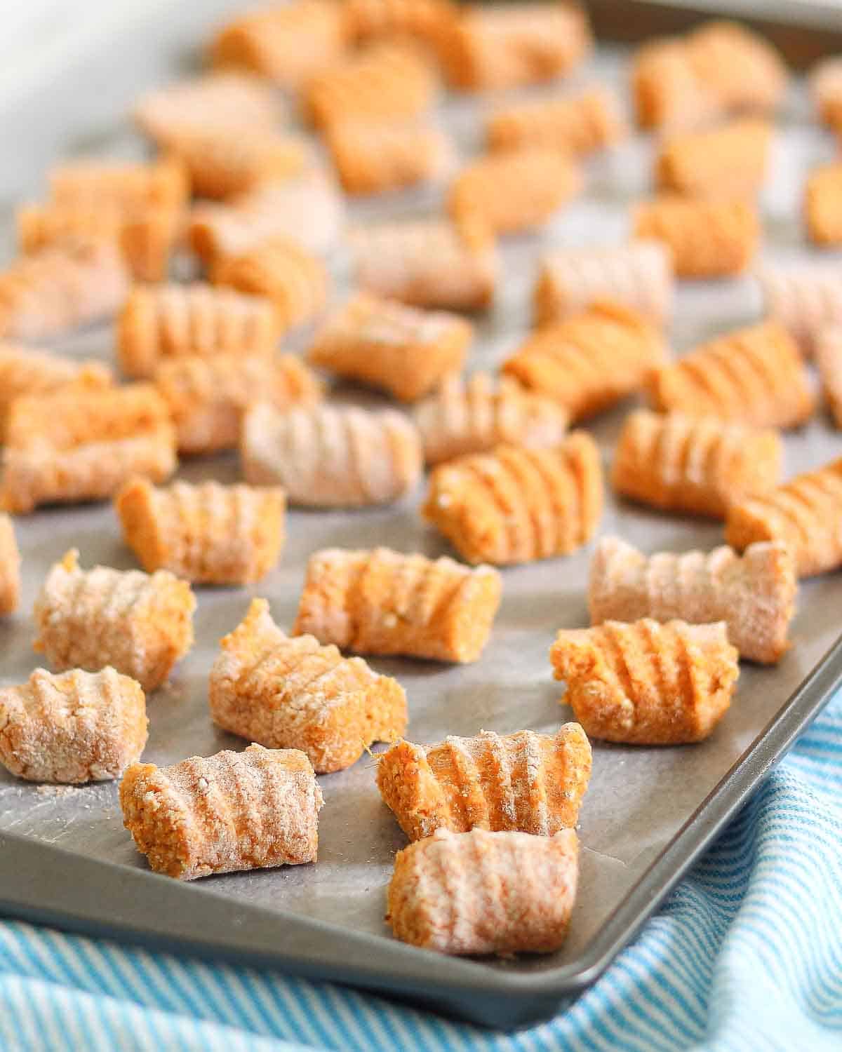 A baking tray filled with uncooked homemade sweet potato gnocchi, lightly dusted with flour. The gnocchi pieces are evenly spaced and have a ridged texture. The tray is on a striped blue cloth.