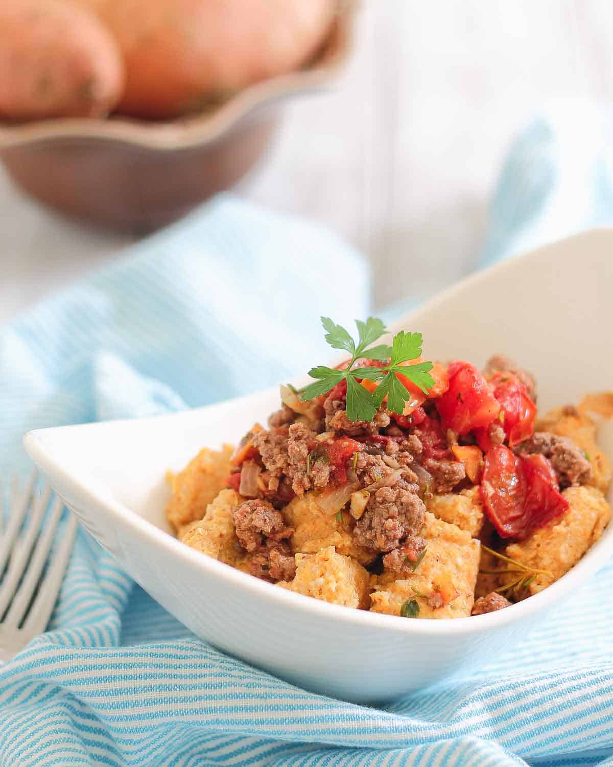A white bowl filled with sweet potato gnocchi topped with cooked ground meat and cherry tomatoes, garnished with parsley. A blue striped cloth and a fork are next to the bowl.