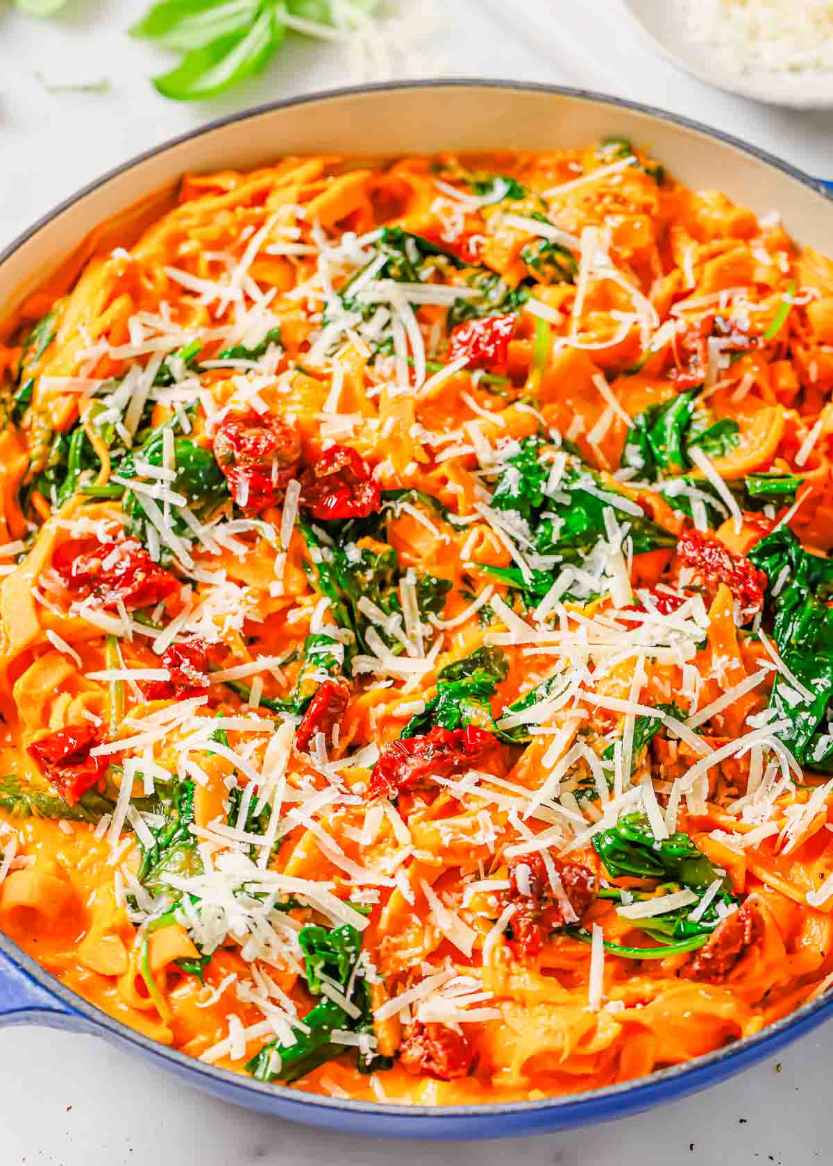 A close-up of a skillet containing creamy sweet potato noodles topped with spinach, sun-dried tomatoes, and shredded parmesan cheese.
