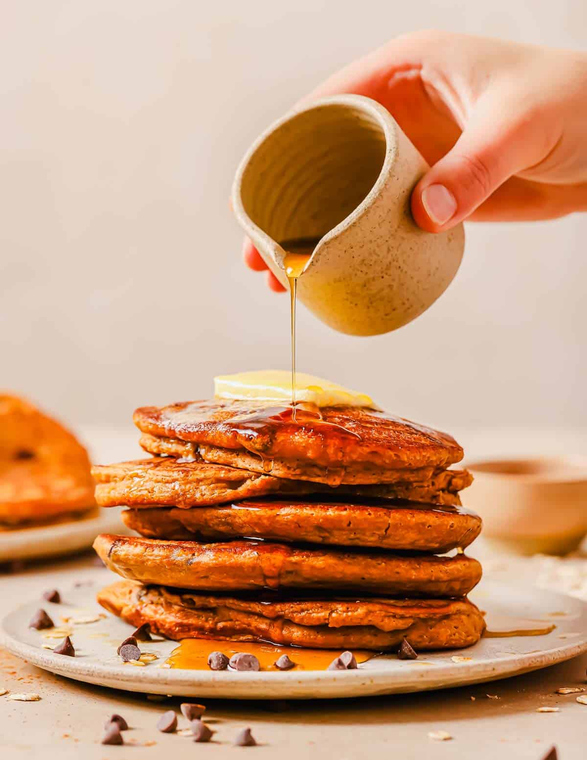 A stack of sweet potato pancakes topped with butter being drizzled with syrup from a small jug held by a hand. Chocolate chips are scattered on the plate and the table.