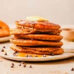 A stack of six pancakes topped with a pat of butter and drizzled with syrup, surrounded by chocolate chips on a beige plate.