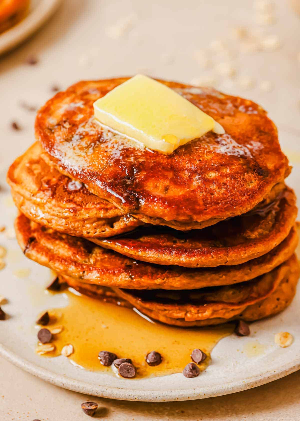 A stack of four sweet potato pancakes topped with a pat of butter and syrup on a plate, surrounded by scattered chocolate chips.
