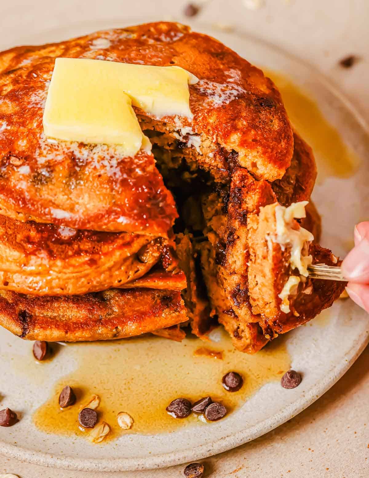 A stack of sweet potato pancakes topped with a pat of butter and syrup, partially cut with a fork taking a bite. Chocolate chips and oats are scattered around the plate.