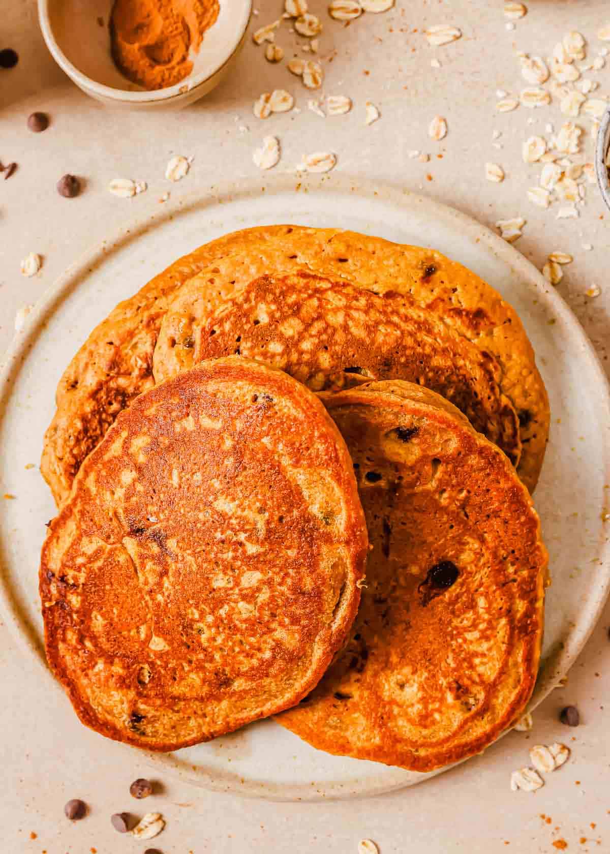 A plate of four golden-brown sweet potato chocolate chip pancakes with visible oats, placed on a light-colored surface scattered with oats and chocolate chips. A small bowl of cinnamon lies above the plate.