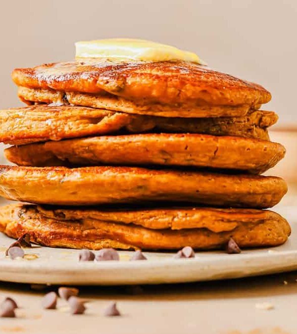 A stack of sweet potato pancakes with chocolate chips topped with a pat of butter on a white plate, surrounded by scattered chocolate chips on a light-colored surface.