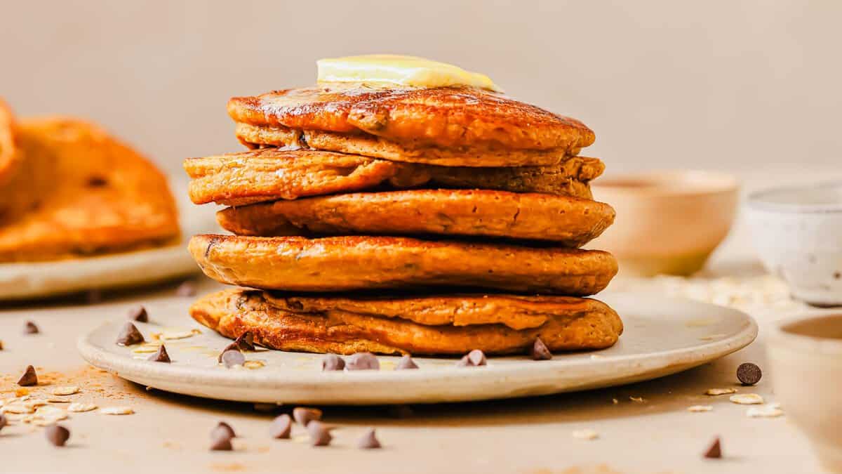 A stack of sweet potato pancakes with chocolate chips topped with a pat of butter on a white plate, surrounded by scattered chocolate chips on a light-colored surface.
