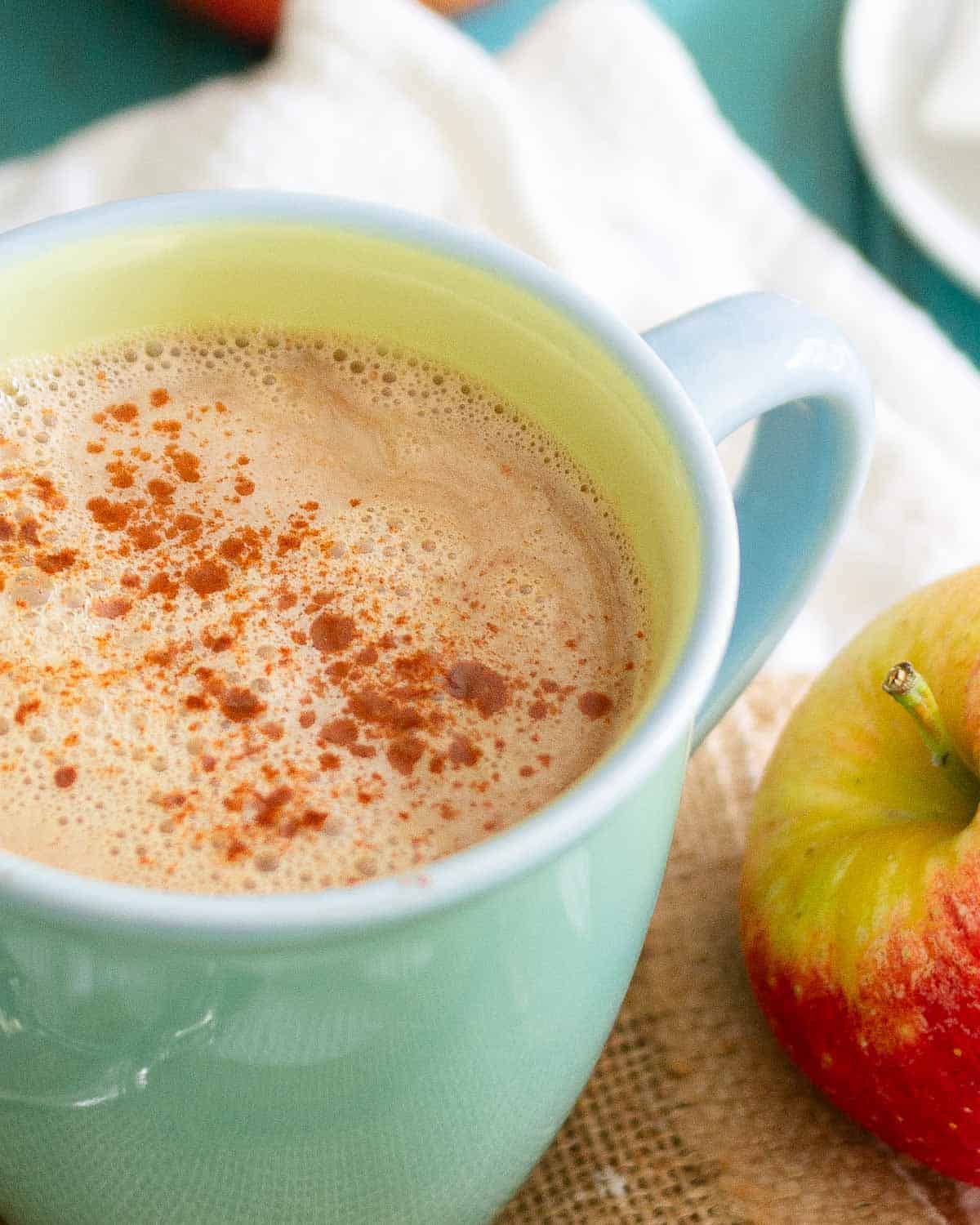 A green mug filled with an apple cinnamon tea latte, topped with a sprinkle of spice, next to a red and yellow apple.
