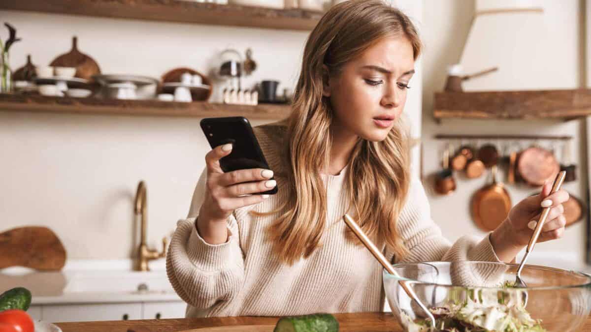 A woman in a kitchen holding a smartphone in one hand while mixing a salad in a glass bowl with chopsticks in the other hand.