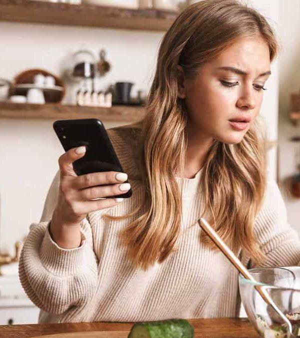 A woman in a kitchen holding a smartphone in one hand while mixing a salad in a glass bowl with chopsticks in the other hand.
