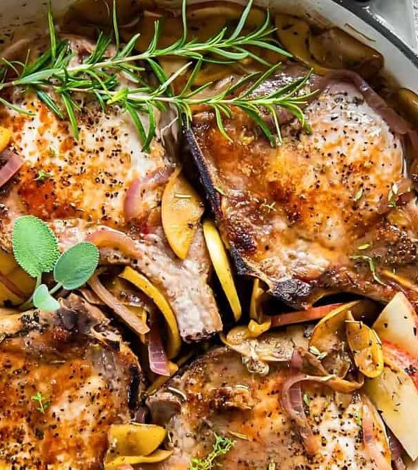 A skillet with seasoned pork chops garnished with fresh rosemary, surrounded by sliced apples and onions. The dish is presented on a woven mat with a linen cloth in the background.