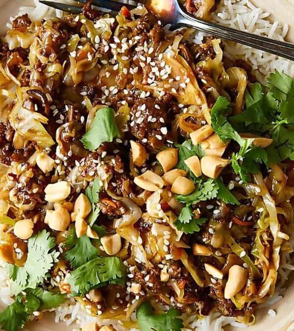 A bowl of rice topped with beef, caramelized onions, peanuts, cilantro, and sesame seeds, with a fork resting on the side.