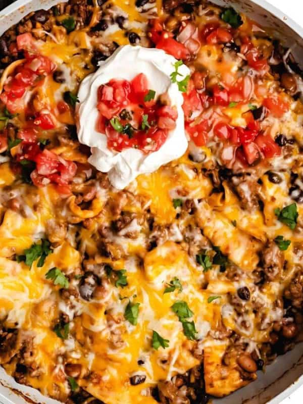 A skillet filled with cheesy taco pasta topped with melted cheese, black beans, diced tomatoes, and a dollop of sour cream. Fresh cilantro is sprinkled on top, adding a touch of green. A wooden spoon is nearby on a white background.