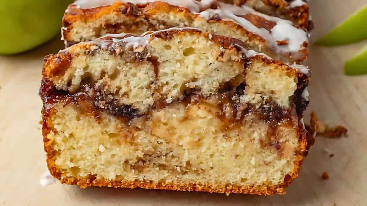 A close-up of a sliced loaf of apple cinnamon bread with a drizzle of icing on top, showing a moist, marbled interior.