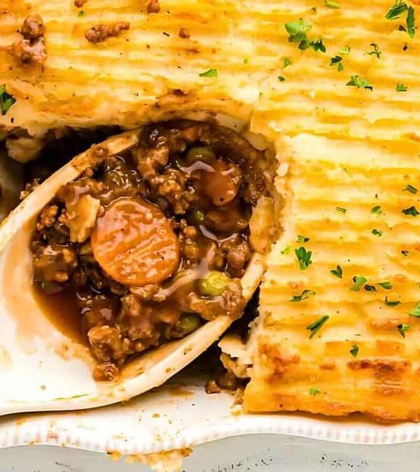 A baked shepherd's pie in a dish with a portion removed, showing the meat and vegetable filling beneath a layer of mashed potatoes. Garnished with chopped parsley.