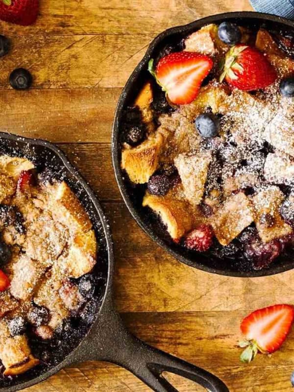 Two cast iron skillets filled with baked blueberry and strawberry bread pudding topped with powdered sugar. A bowl of blueberries and a cup of coffee sit nearby on a rustic wooden table.