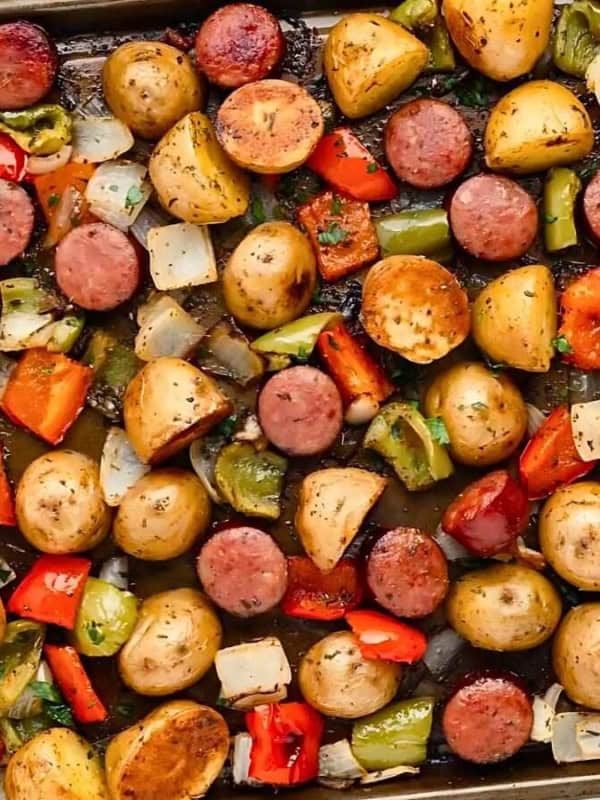 A sheet pan with roasted sausage slices, baby potatoes, and mixed vegetables, including red and green bell peppers and onions, garnished with herbs. The food is evenly spread and appears freshly baked.