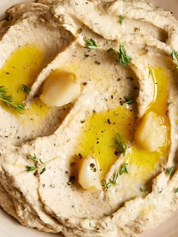 A bowl of hummus garnished with olive oil, roasted garlic cloves, and fresh herbs. The smooth texture is enhanced by a sprinkle of black pepper, set on a blue wooden surface.