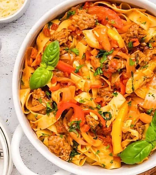 A white pot filled with tagliatelle pasta topped with a tomato-based sauce, bell peppers, and fresh basil leaves. It is surrounded by plates, a wooden spoon, silver cutlery, grated cheese, chopped herbs, and salt and pepper shakers.