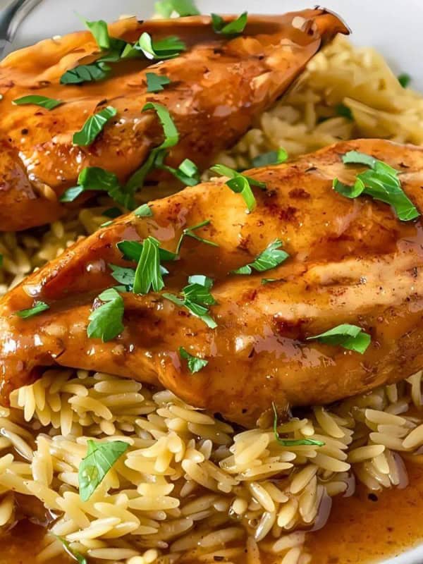 A white plate with two pieces of glazed chicken breast, garnished with parsley, served on a bed of rice or orzo, accompanied by a fork and spoon. The chicken is covered in a glossy sauce.