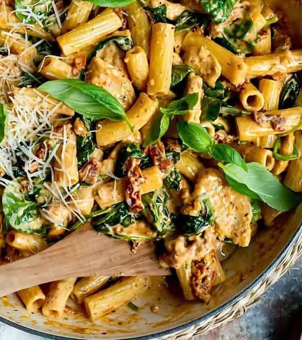 A skillet filled with creamy rigatoni pasta featuring pieces of chicken, spinach, and sun-dried tomatoes. The dish is garnished with fresh basil leaves and a sprinkle of grated cheese, accompanied by a wooden spoon.