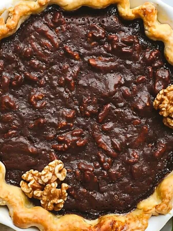 A freshly baked pie with a golden, crimped crust filled with a dark chocolate and nut mixture, garnished with whole walnuts. The pie is set on a marble surface next to a folded napkin, scattered walnuts, and a rolling pin dusted with flour.
