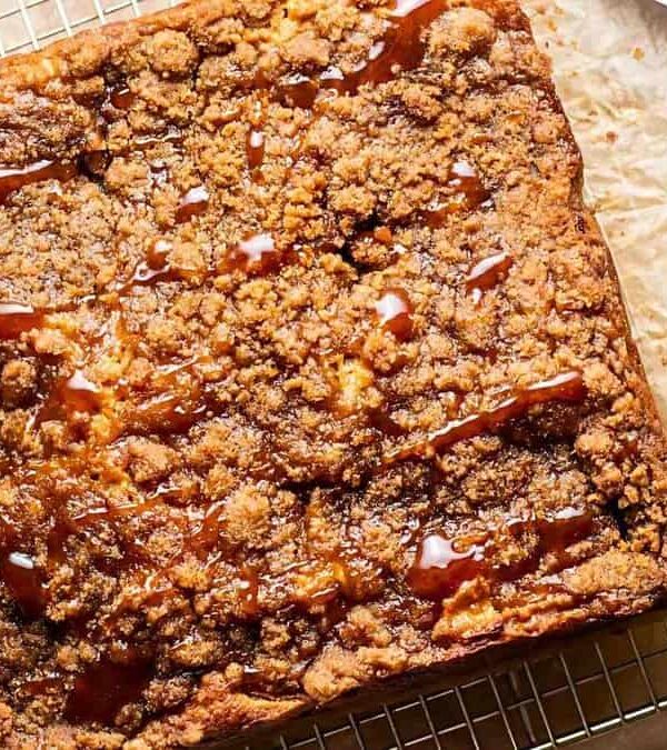 A square crumb cake with caramel drizzle sits on a wire cooling rack with parchment paper. A cup of caramel sauce and a spoon with caramel are beside it.