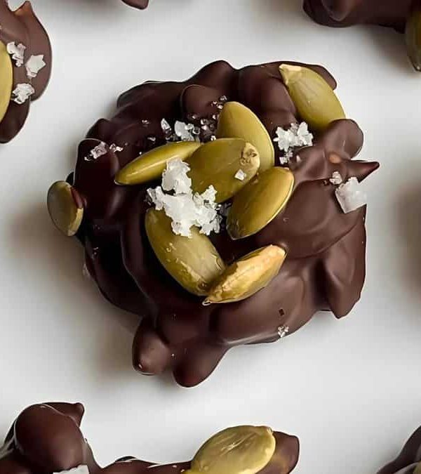 Clusters of dark chocolate are topped with pumpkin seeds and sprinkled with coarse sea salt, arranged on a white background.