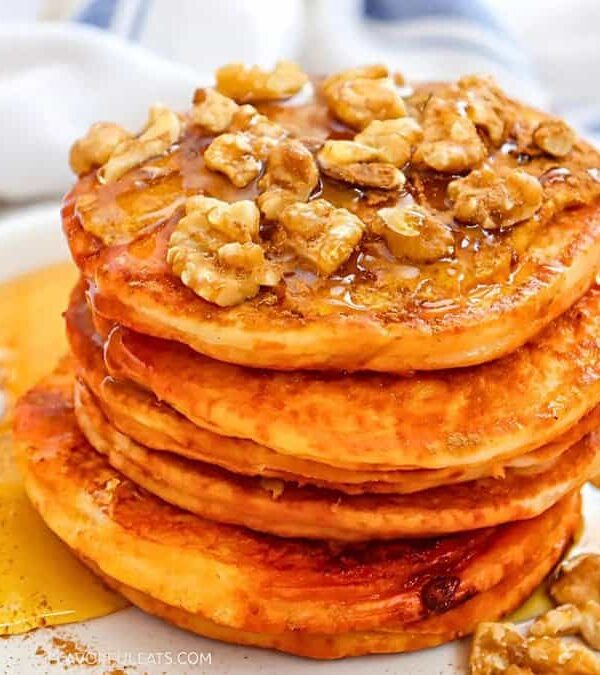 A stack of pancakes topped with chopped walnuts and drizzled with syrup sits on a white plate. A white cloth napkin and fork are in the background, creating a cozy breakfast scene.