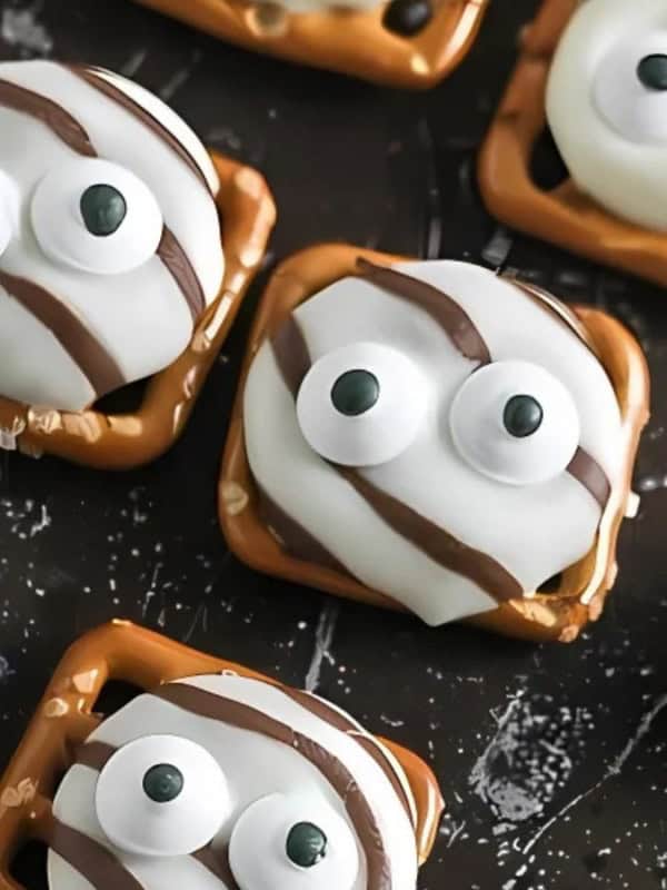 Pretzel squares topped with candy-coated chocolates designed to look like eyes, drizzled with white and dark chocolate. The treats are arranged on a black surface dusted with flour or powdered sugar.
