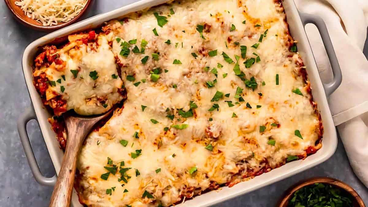 A baked lasagna in a rectangular dish, topped with melted cheese and fresh parsley. A wooden spoon is serving a piece, revealing layers of pasta, meat, and sauce. Bowls of grated cheese and chopped herbs are nearby.