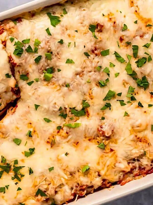 A baked lasagna in a rectangular dish, topped with melted cheese and fresh parsley. A wooden spoon is serving a piece, revealing layers of pasta, meat, and sauce. Bowls of grated cheese and chopped herbs are nearby.