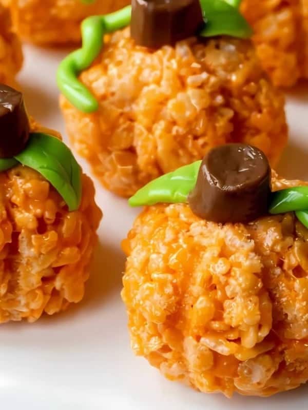 Close-up of pumpkin-shaped treats made from orange rice cereal, topped with chocolate stems and green icing leaves, arranged on a white plate.