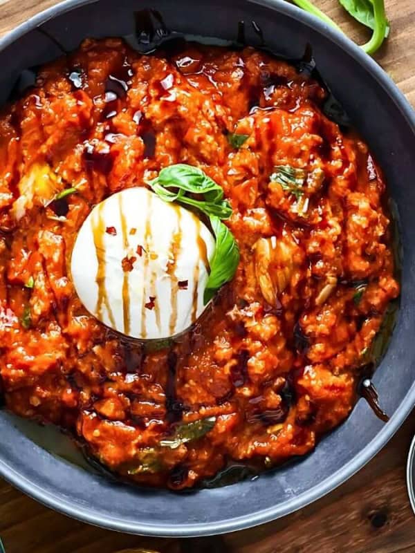 A black bowl with a rich, red tomato eggplant stew topped with a ball of burrata cheese, garnished with basil. Surrounded by bread, salt, peppercorns, and fresh burrata, set on a wooden table with a spoon and napkin.