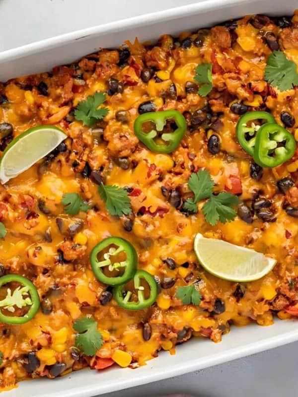 A baked Tex-Mex casserole in a white dish, topped with melted cheese, jalapeño slices, lime wedges, and cilantro. Surrounding are forks, halved jalapeños, lime wedges, fresh cilantro, and a small bowl of sour cream.