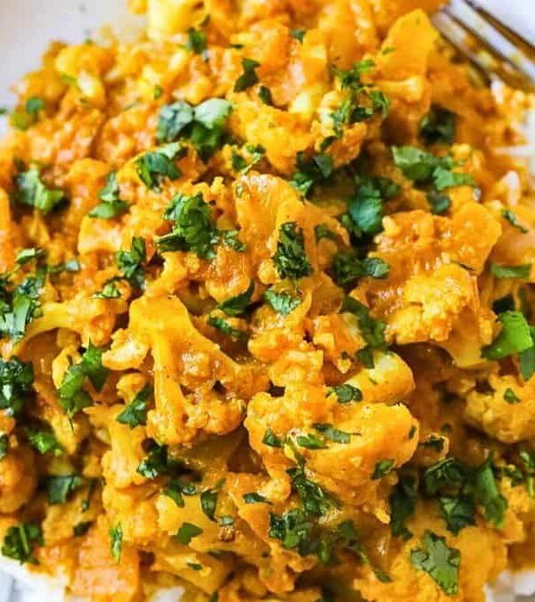 A white bowl filled with a vibrant curry, featuring golden cauliflower and diced vegetables garnished with chopped cilantro. The dish is served over a bed of white rice, accompanied by a fork on the side.