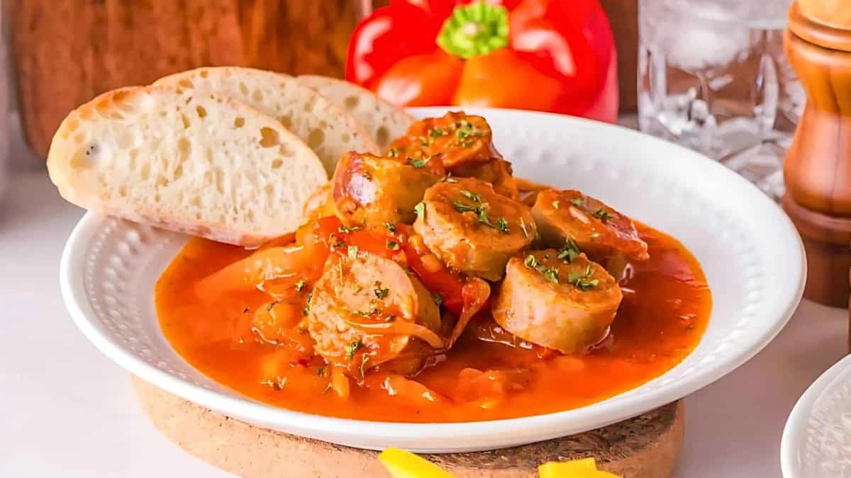 A white plate holds sausage slices in a rich tomato-based sauce, garnished with herbs, beside sliced bread. A red bell pepper and a glass of water are in the background, along with a pepper grinder.