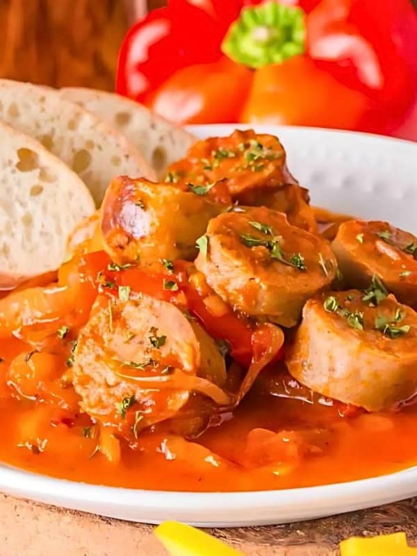 A white plate holds sausage slices in a rich tomato-based sauce, garnished with herbs, beside sliced bread. A red bell pepper and a glass of water are in the background, along with a pepper grinder.