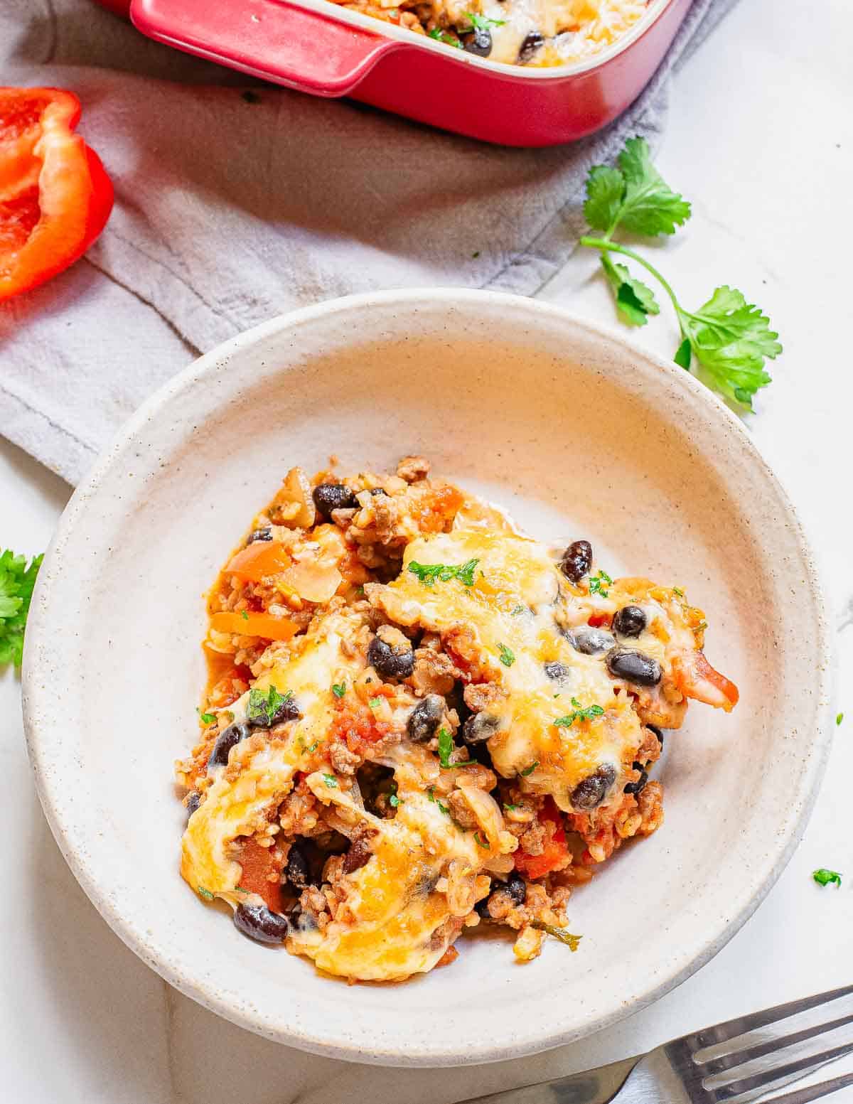 A bowl of cheesy Mexican beef casserole with black beans and vegetables. A red casserole dish and a slice of red bell pepper are in the background.