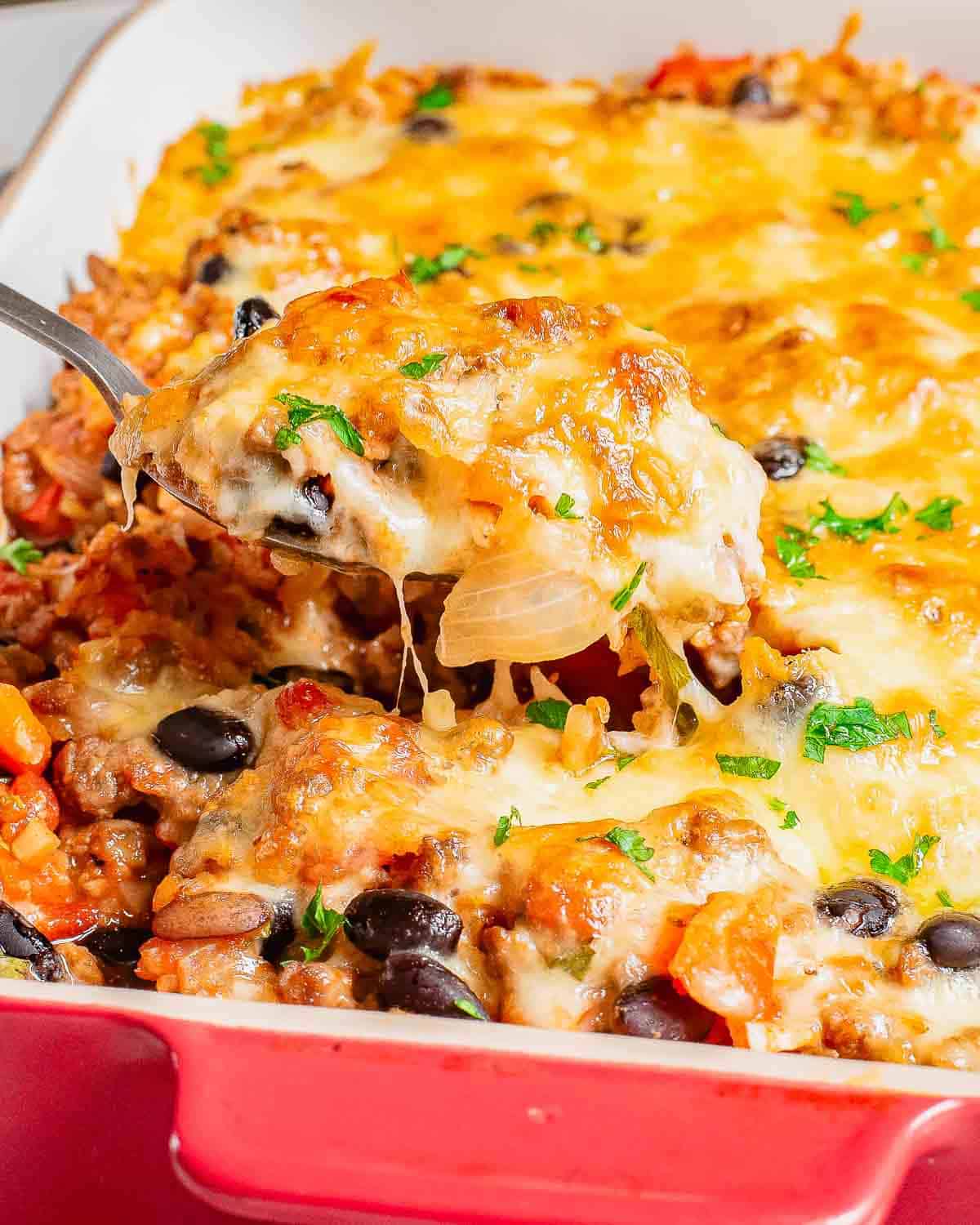 A spoonful of cheesy ground beef casserole with black beans, rice, and vegetables being served from a red baking dish.