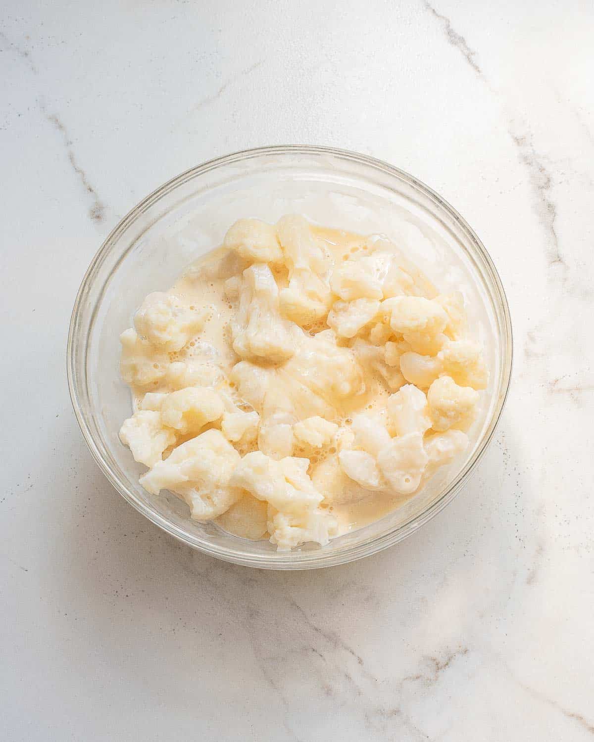 A glass bowl filled with cauliflower florets coated in a creamy white batter sits on a white marble surface.