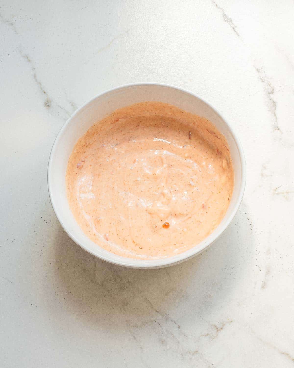 A white bowl filled with bang bang sauce sits on a white marble surface.