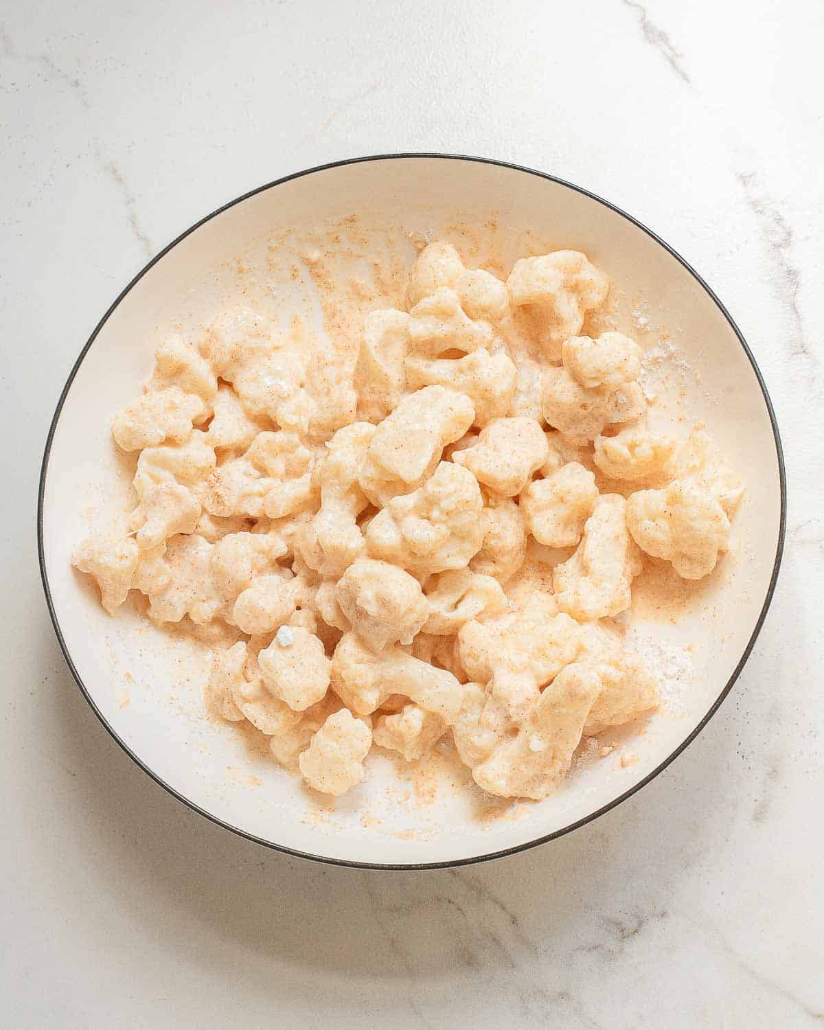 A white plate filled with pieces of cauliflower coated in a batter and light dusting of cornstarch, placed on a white marble surface.